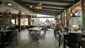 A spacious restaurant with high ceilings, exposed beams, and large windows. Tables are arranged for dining, and there are hanging light fixtures.