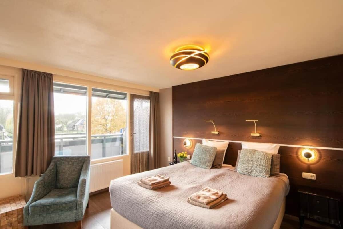 A hotel room with a large bed, a chair, a view of the outside, and a modern ceiling light fixture.