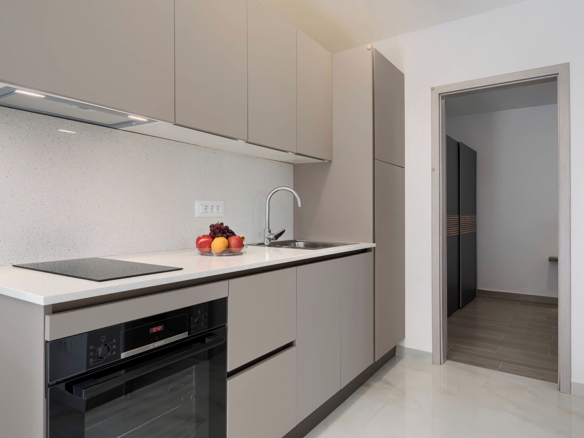 A modern kitchen with a built-in oven, a stainless steel sink, and a fruit bowl on the countertop.