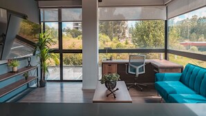 Une chambre moderne équipée d’un bureau, d’une chaise, d’un canapé et de grandes fenêtres donnant sur un paysage verdoyant et des bâtiments.
