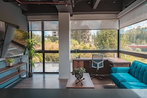 Ein modernes Zimmer mit Schreibtisch, Stuhl, Sofa und großen Fenstern mit Blick auf Grünflächen und Gebäude.