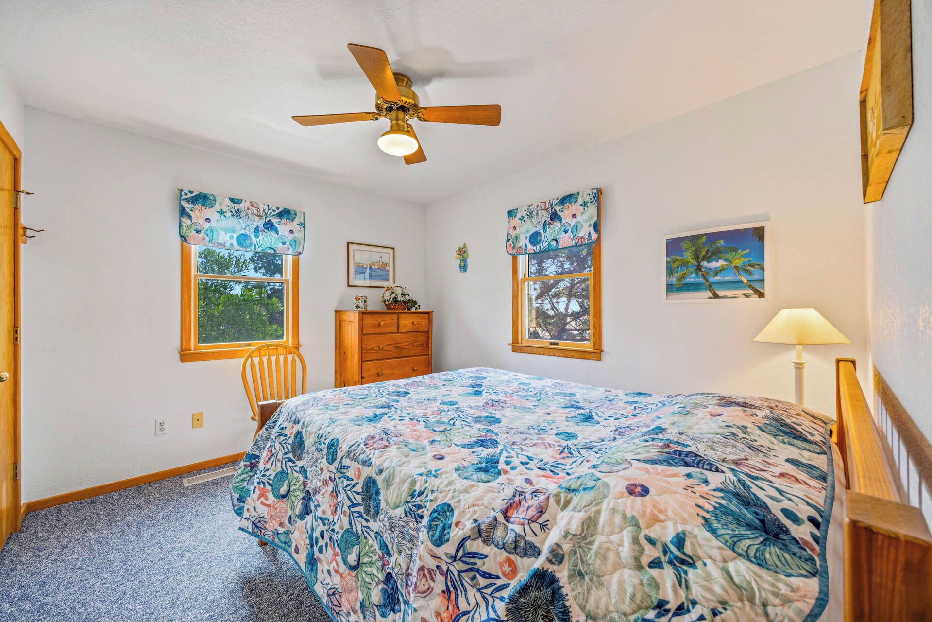 A bedroom with a bed, dresser, chair, ceiling fan, windows, and artwork.