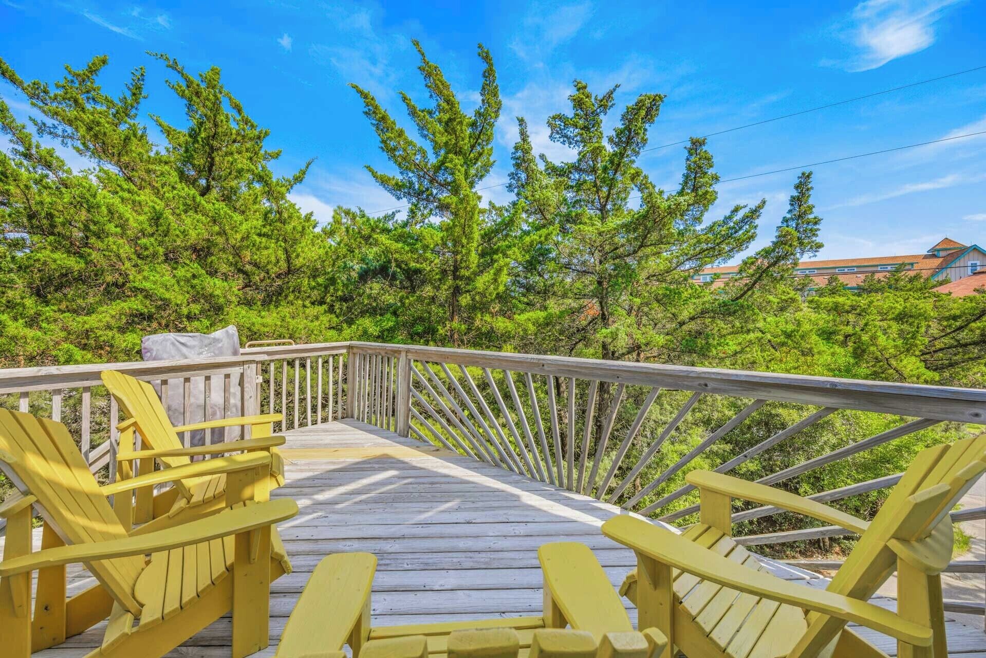 Une terrasse en bois avec des fauteuils Adirondack jaunes, entourée d’arbres et avec un bâtiment visible au loin.