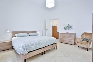 Ein Schlafzimmer mit Bett, Nachttischen, einem Schminktisch, einem Sessel und einer Tür zu einem weiteren Raum.