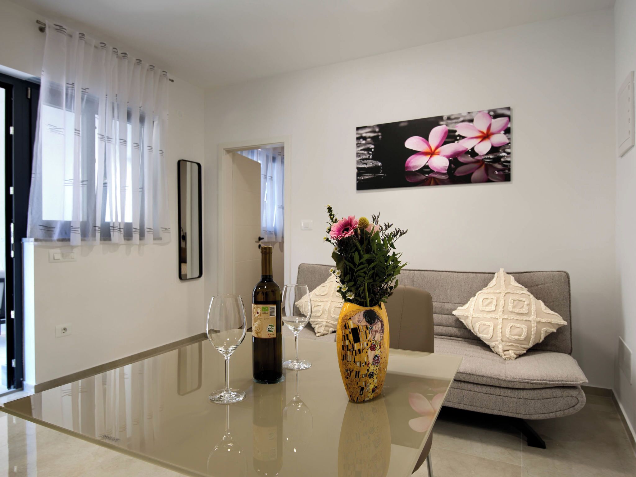 A living room with a glass table, a sofa, a vase with flowers, a wine bottle, and a framed picture on the wall.