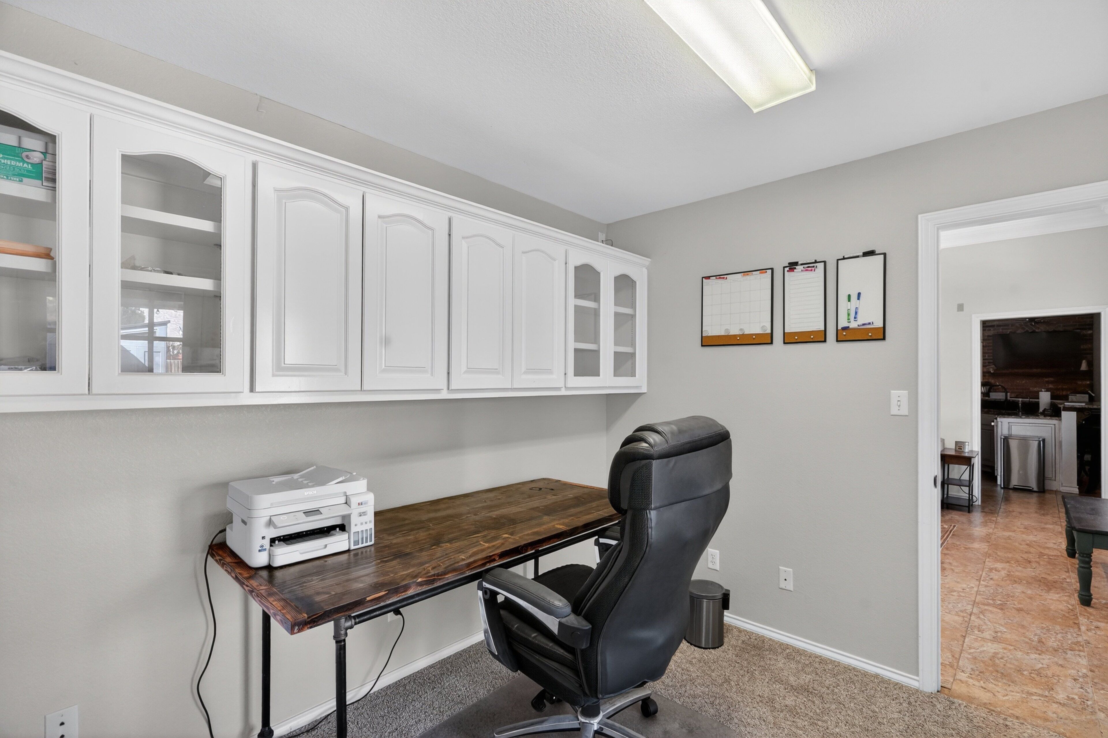 A home office with a desk, chair, cabinets, and a kitchen in the background.