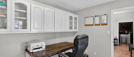 A home office with a desk, chair, cabinets, and a kitchen in the background.