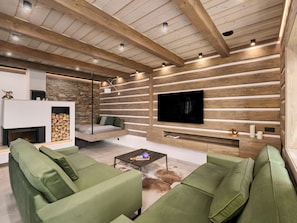 Ein modernes Wohnzimmer mit Kamin aus Stein, grünen Sofas, einem Flachbildfernseher an einer Holzwand und einer Decke mit freiliegenden Balken.