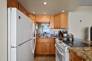 A compact kitchen with wooden cabinets, a white refrigerator, a stove, and a granite countertop. - Kihei Beach, #108 1 Bedroom Condo (Kihei)