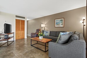 A living room with a sofa, armchair, coffee table, TV, and a painting on the wall. - Kihei Beach, #108 1 Bedroom Condo (Kihei)