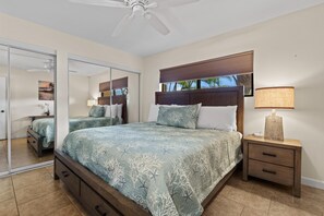 A bedroom with a bed, a nightstand, a mirror, and a ceiling fan. - Kihei Beach, #108 1 Bedroom Condo (Kihei)