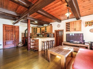Un interior rústico con techo de madera, una zona de cocina con armarios de madera, un taburete de bar, una mesa de comedor de madera y un televisor.
