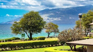 A coastal view with a sign for 'Kamehameha II,' a grassy area with trees, and a distant mountain range. - Hale Kamaole 271- Paradise Found (Kihei)