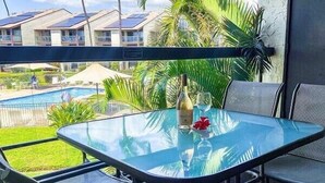 A glass table with chairs, a bottle, and a glass on it, overlooking a swimming pool and buildings. - Hale Kamaole 271- Paradise Found (Kihei)