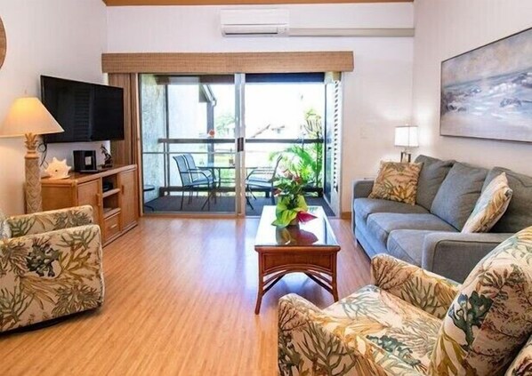 Ein Wohnzimmer mit einer Couch, einem Couchtisch und einem Fernseher. Eine Glastür führt zu einem Außenbereich mit Tisch und Stühlen.