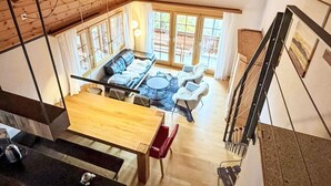Ein Loft-Zimmer mit hoher Decke, Holzbalken, einem Essbereich mit Tisch und Stühlen, einer Sofagarnitur und einem großen Fenster mit Vorhängen.