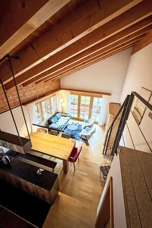 Ein Loft-Zimmer mit hoher Decke, Holzbalken, einem Essbereich mit Tisch und Stühlen, einer Sofagarnitur und einem großen Fenster mit Vorhängen.