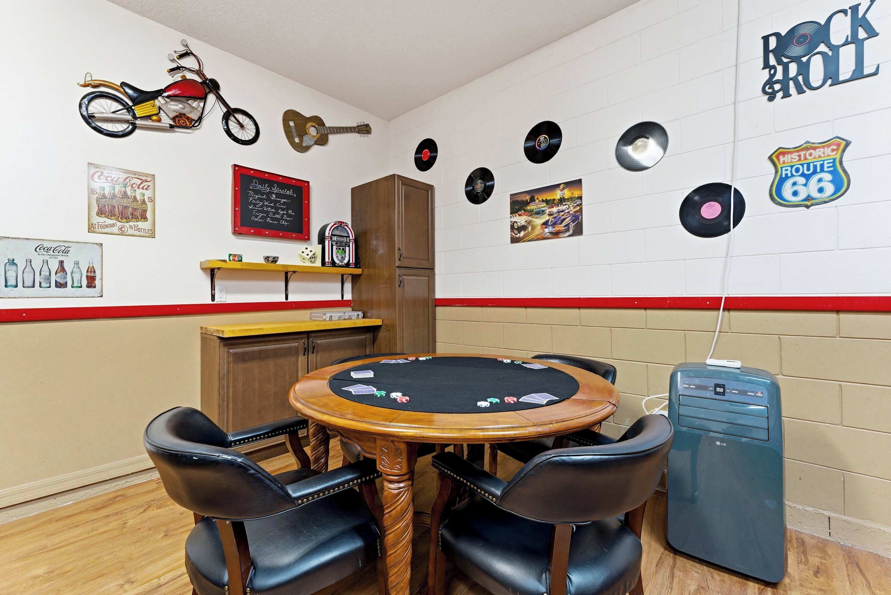 Une pièce comprenant une table de poker, des chaises, un juke-box et des éléments de décoration murale, notamment une moto, une guitare et des disques vinyle.