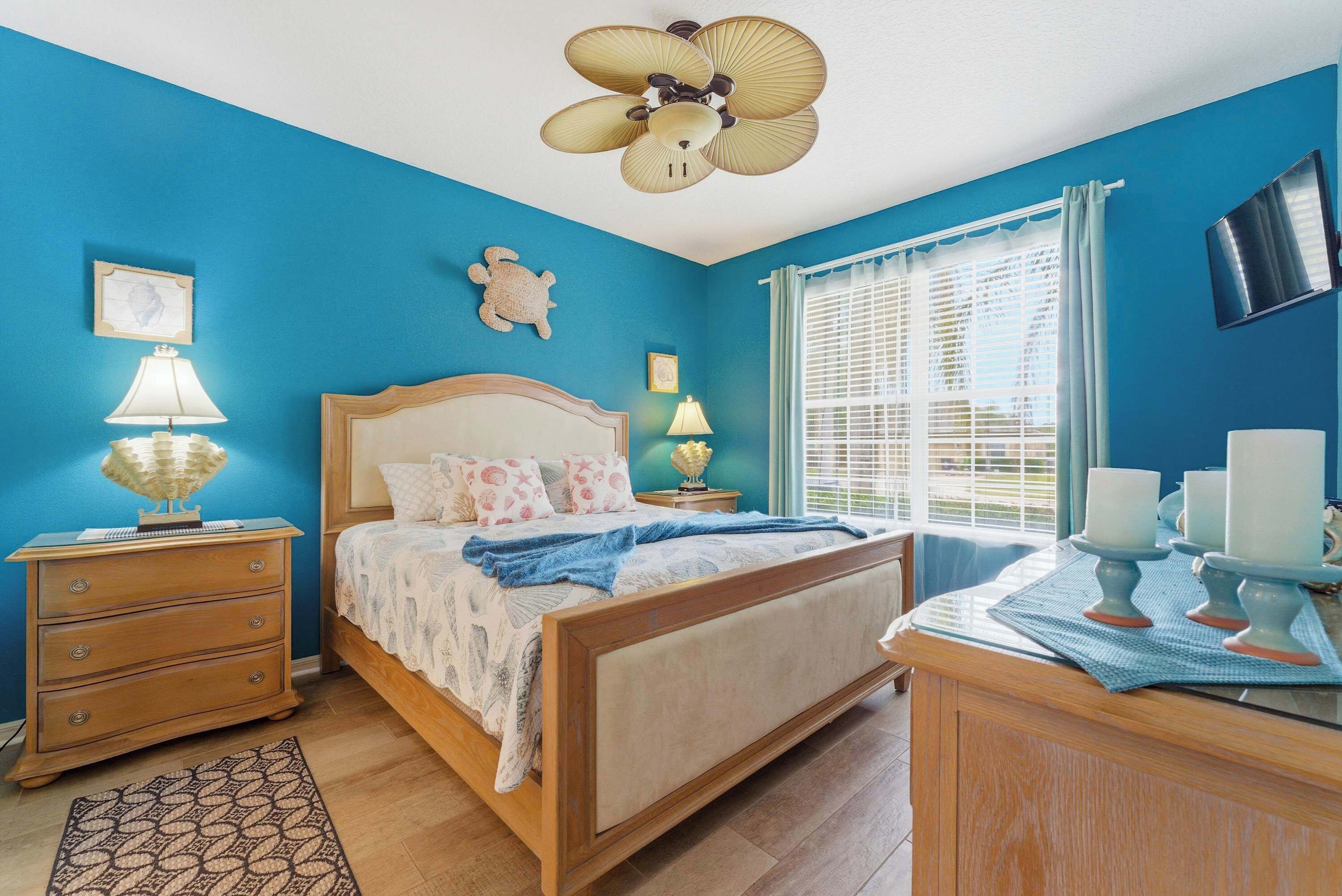 A bedroom with a bed, bedside tables, a ceiling fan, a window with curtains, and a wall-mounted TV.
