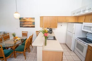 A compact kitchen with wooden cabinets, a white countertop, a dining table with chairs, a coffee maker, and a refrigerator. - Island Retreat - Two bedroom Two bathroom Hale Kamaole 329 (Kihei)