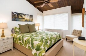 Ein Schlafzimmer mit Bett, Nachttischen, einem Sessel, Schreibtisch, Deckenventilator und einem Fenster mit Jalousien.