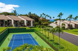 A tennis court surrounded by hotel buildings with balconies, palm trees, and a clear sky. - Island Retreat - Two bedroom Two bathroom Hale Kamaole 329 (Kihei)