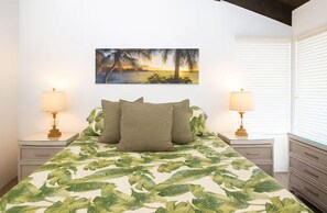 A bedroom with a bed, two bedside lamps, a nightstand, a dresser, and a painting of a sunset over water. - Island Retreat - Two bedroom Two bathroom Hale Kamaole 329 (Kihei)