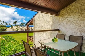 A balcony with a table and chairs, a view of a golf course, and a building with a stone facade. - Island Retreat - Two bedroom Two bathroom Hale Kamaole 329 (Kihei)