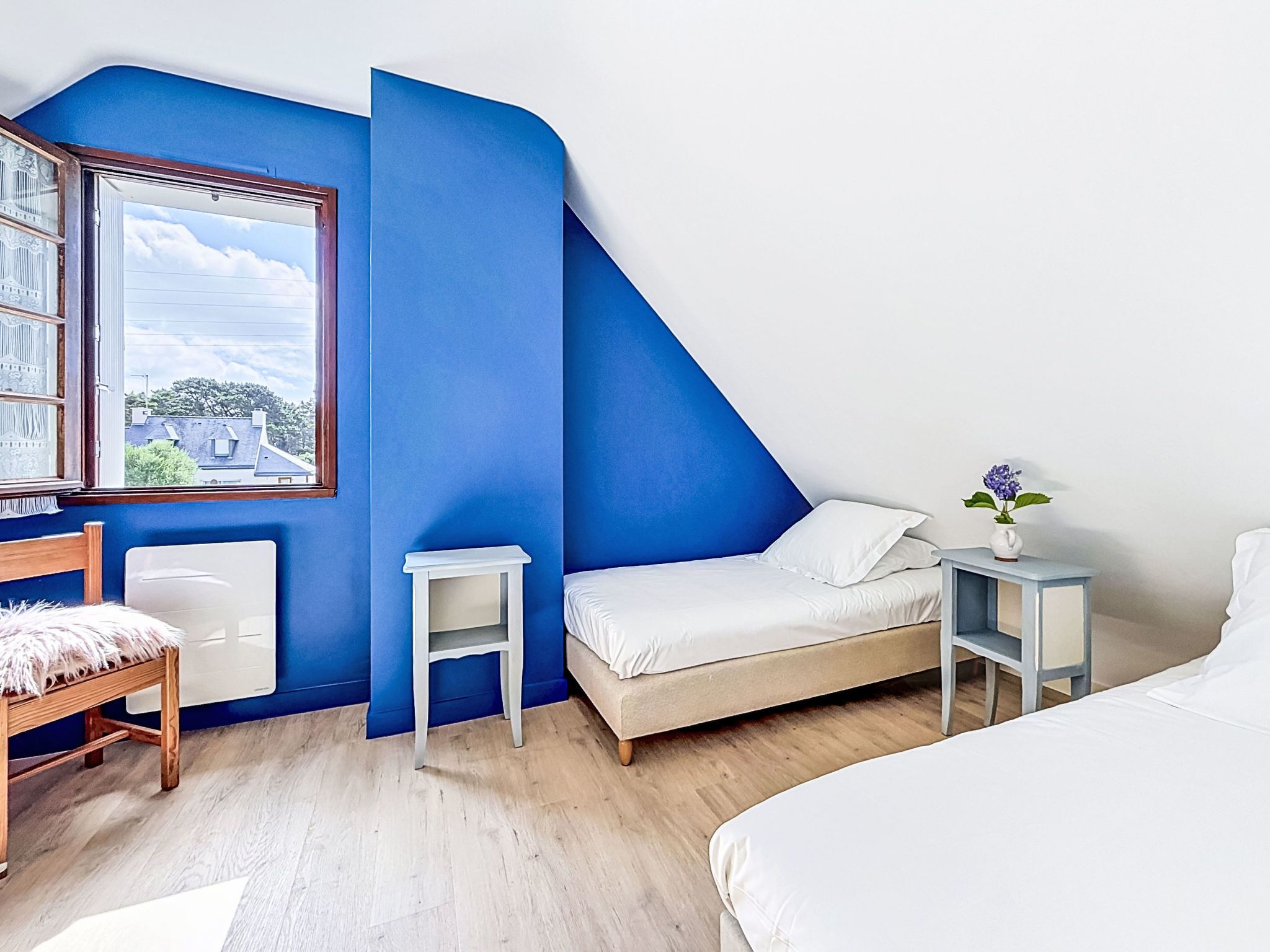 Ein Schlafzimmer mit schräg verlaufender Decke, einem Fenster, einem Bett, einem Nachttisch mit Vase und einem Sessel.