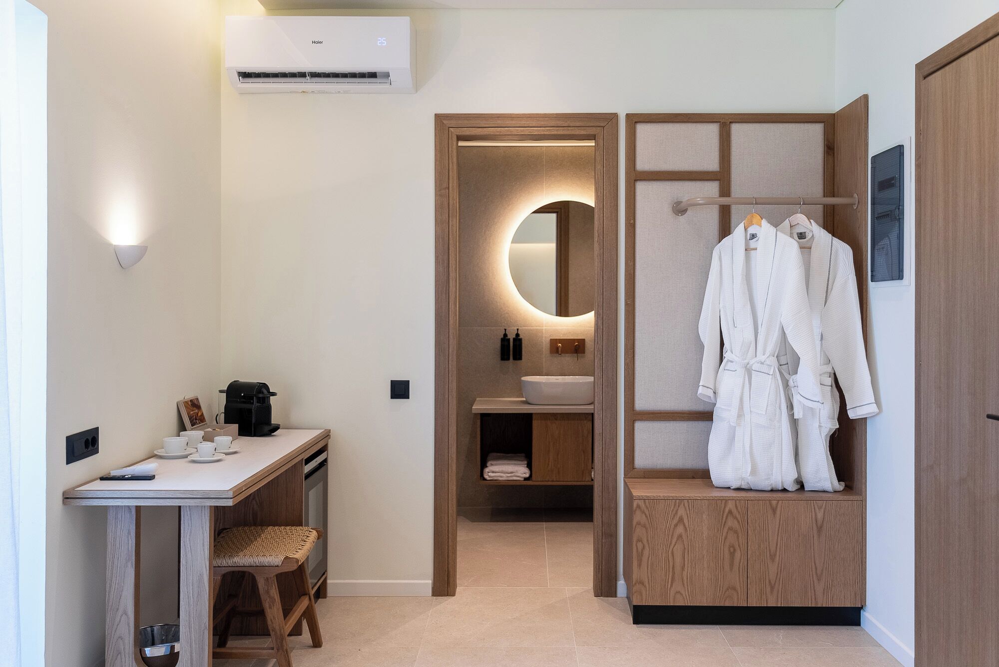 A compact hotel room with a desk, coffee machine, and a mirror with a circular light.