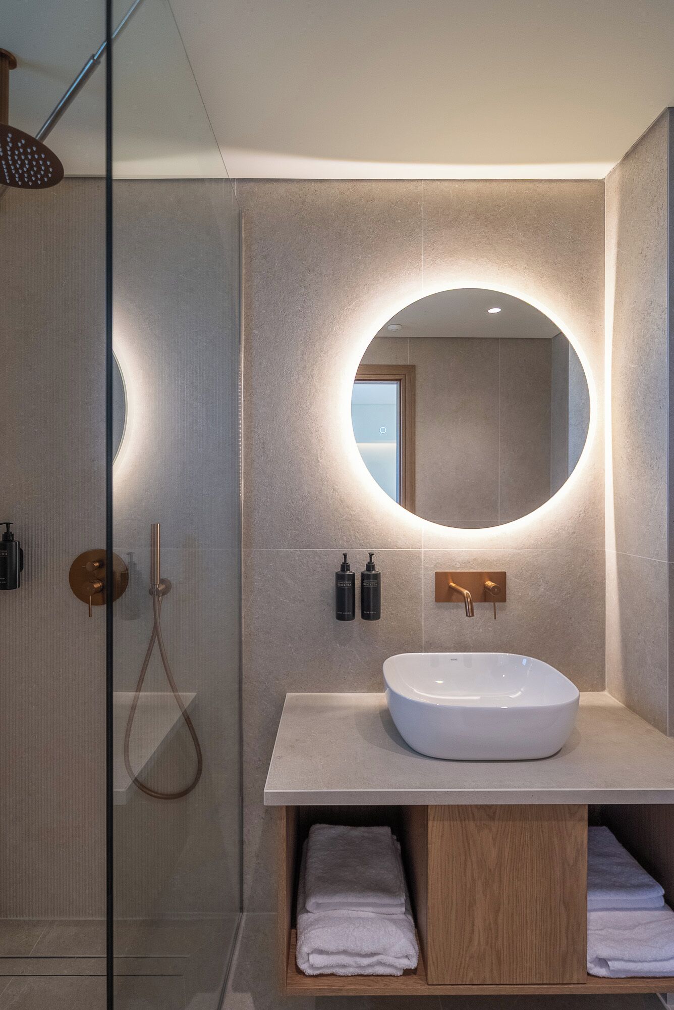 Un baño moderno con cabina de ducha de vidrio, un espejo redondo, un lavabo blanco y toallas dobladas con cuidado.