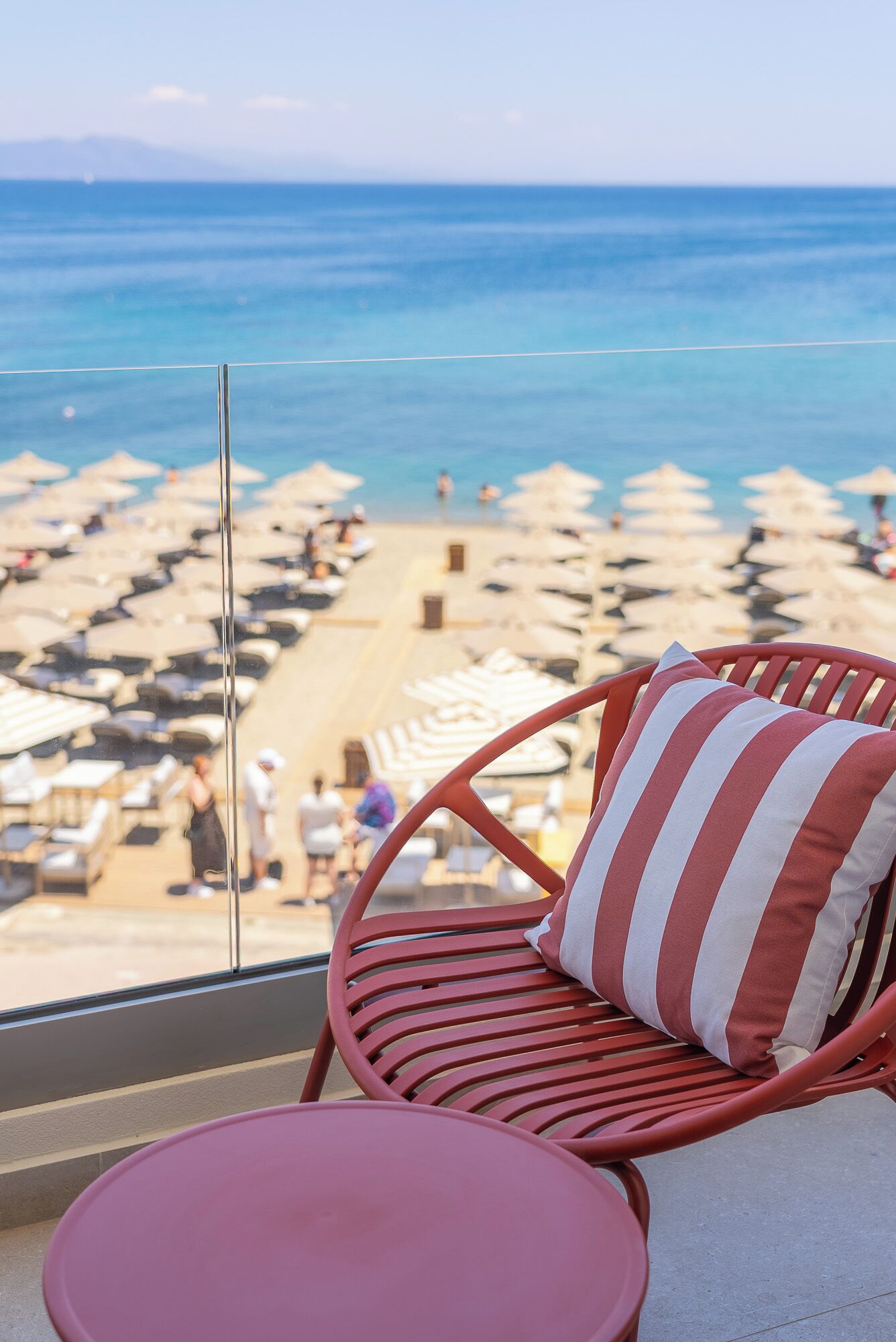Uma cadeira de vime vermelha com almofada listrada, uma mesa rosa e vista para uma praia com guarda-sóis e pessoas.