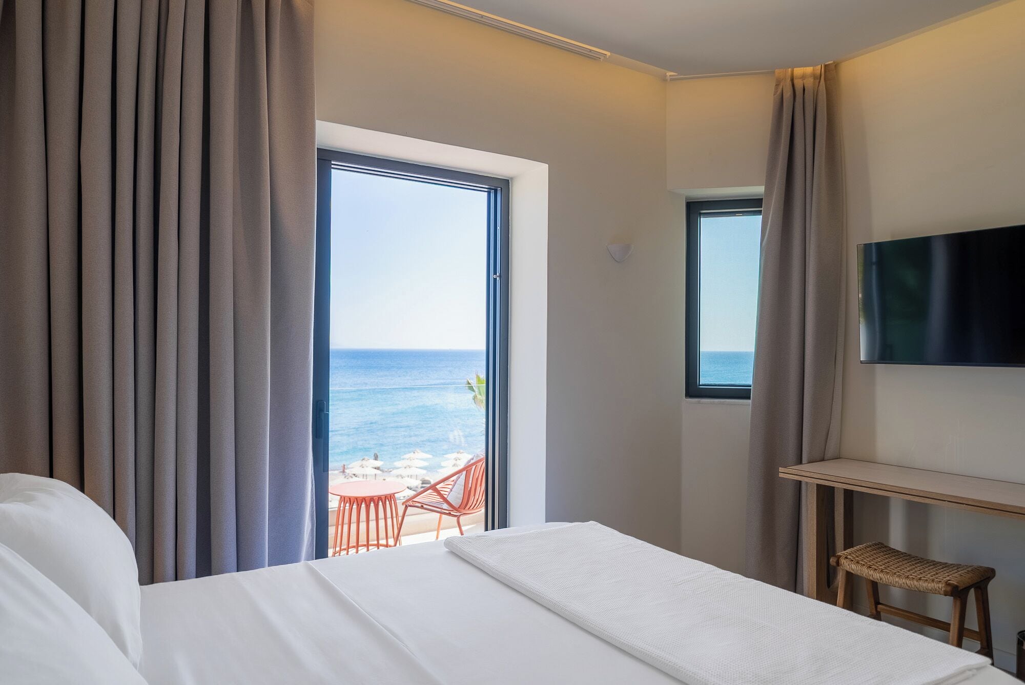 Ein Hotelzimmer mit Bett, Fernseher, kleinem Tisch, Korbsessel und einem Balkon mit Meerblick.