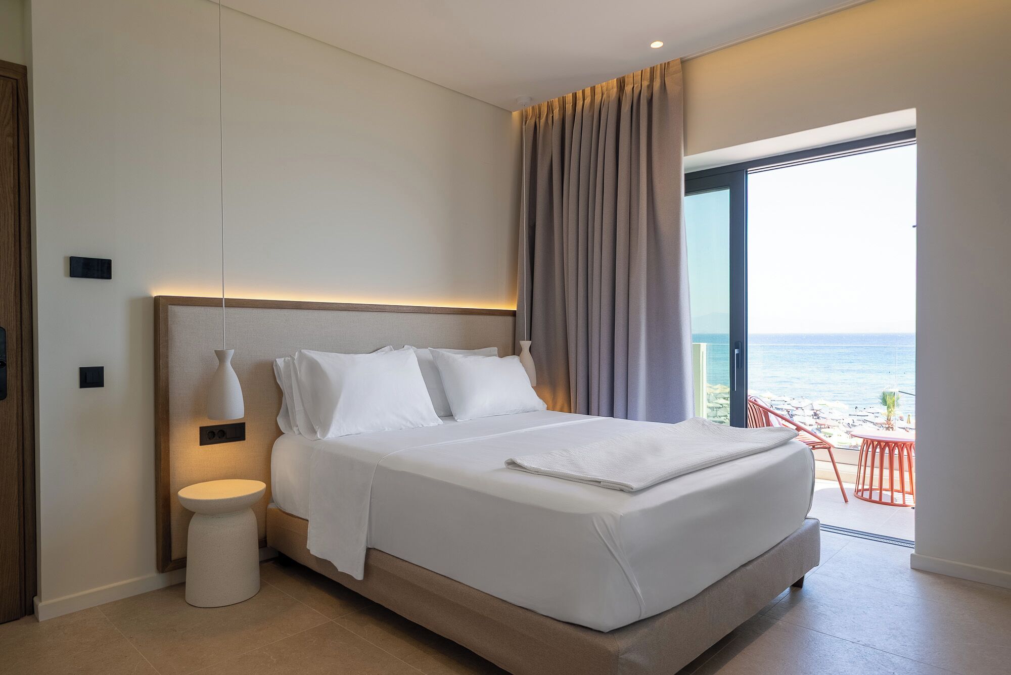 A hotel room with a large bed, a bedside table, a modern lamp, and a view of the beach.