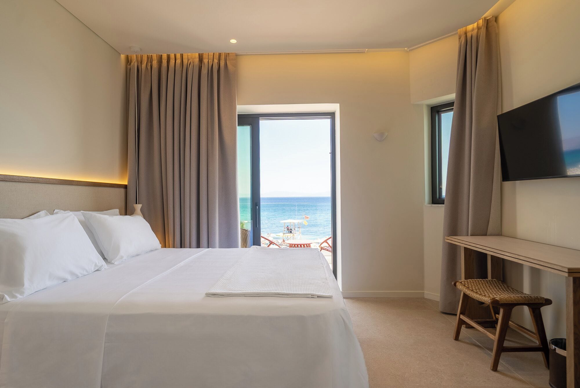 A hotel room with a bed, a desk, a chair, a TV, and a view of the sea.