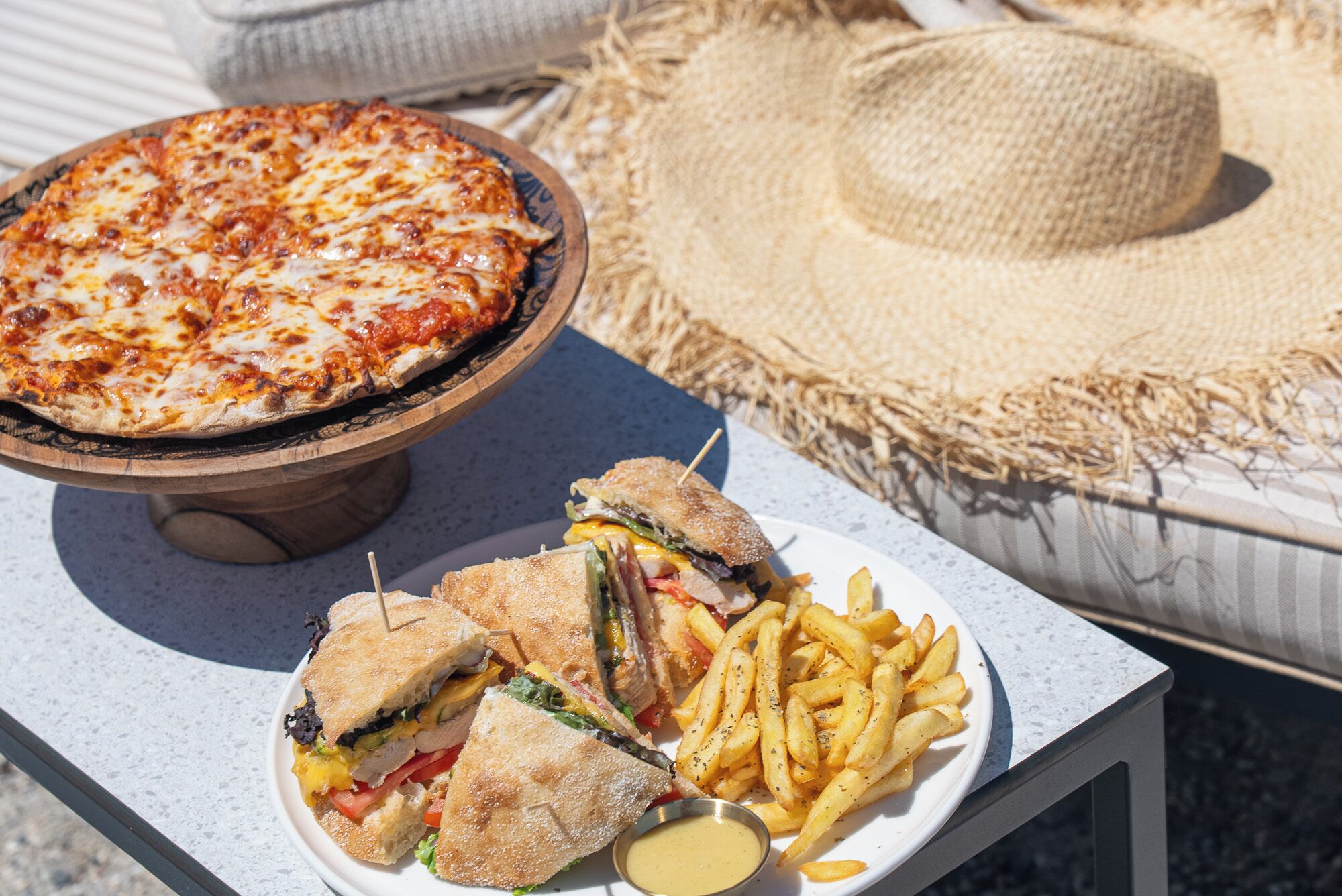 Pizza op een houten standaard, een bord met een sandwich en frietjes, en een strooien hoed op een bank.