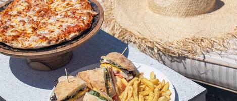Pizza op een houten standaard, een bord met een sandwich en frietjes, en een strooien hoed op een bank.