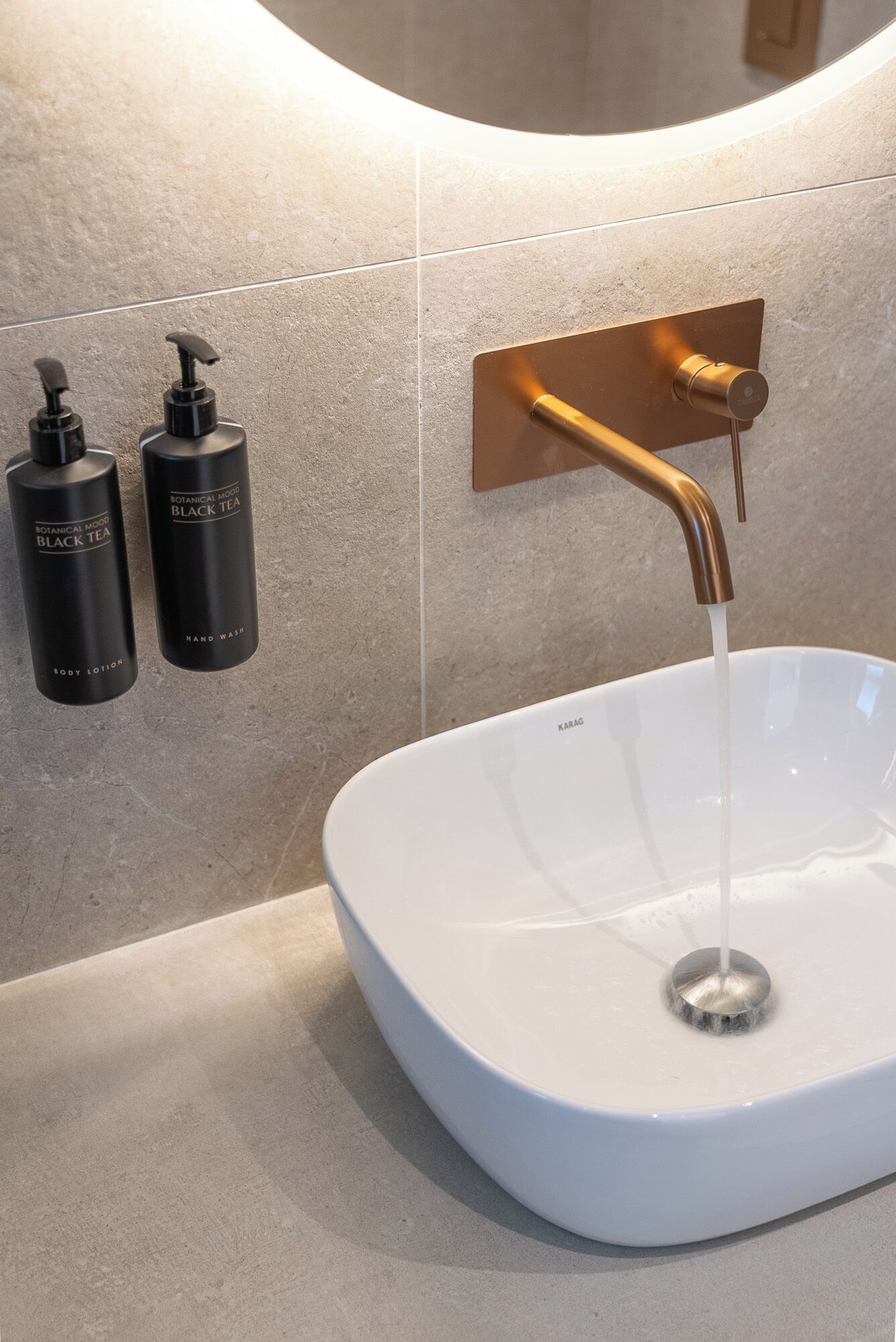 Un lavamanos blanco con un grifo moderno, dos botellas negras etiquetadas como 'Loción para el cuerpo Té Negro Botanical Mood' y 'Jabón de manos', y una etiqueta 'Karag' en el lavamanos.