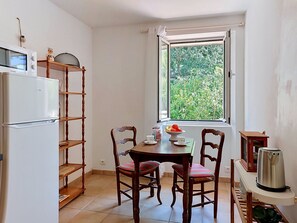 Een functionele keuken met een koelkast, houten planken, een eettafel met stoelen, een raam met uitzicht op groen en een waterkoker op een bijzettafeltje.