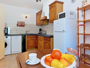 A kitchen with wooden cabinets, a microwave, a refrigerator, a washing machine, and a dining table with a fruit bowl and a cup. - Sainte Marguerite by Interhome (Saint-Tropez)