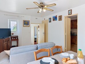 Een kamer met een bank, eettafel, stoelen, een televisie, een plafondventilator en een keukenhoek.