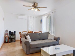 A living room with a grey sofa, a white coffee table, a ceiling fan, a bookshelf, and a dining area with chairs and a table. - Sainte Marguerite by Interhome (Saint-Tropez)