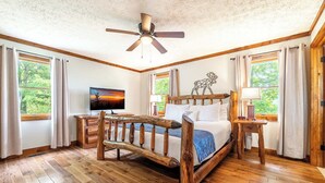 Ein Bett mit Holzkopfende, Deckenventilator, Fernseher und ein Nachttisch mit Lampe in einem Zimmer mit Holzböden und großen Fenstern.