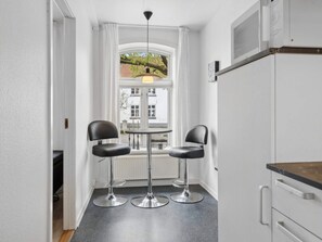 Een kleine keuken met twee zwarte barkrukken, een ronde tafel en een raam met uitzicht op een gebouw en bomen.