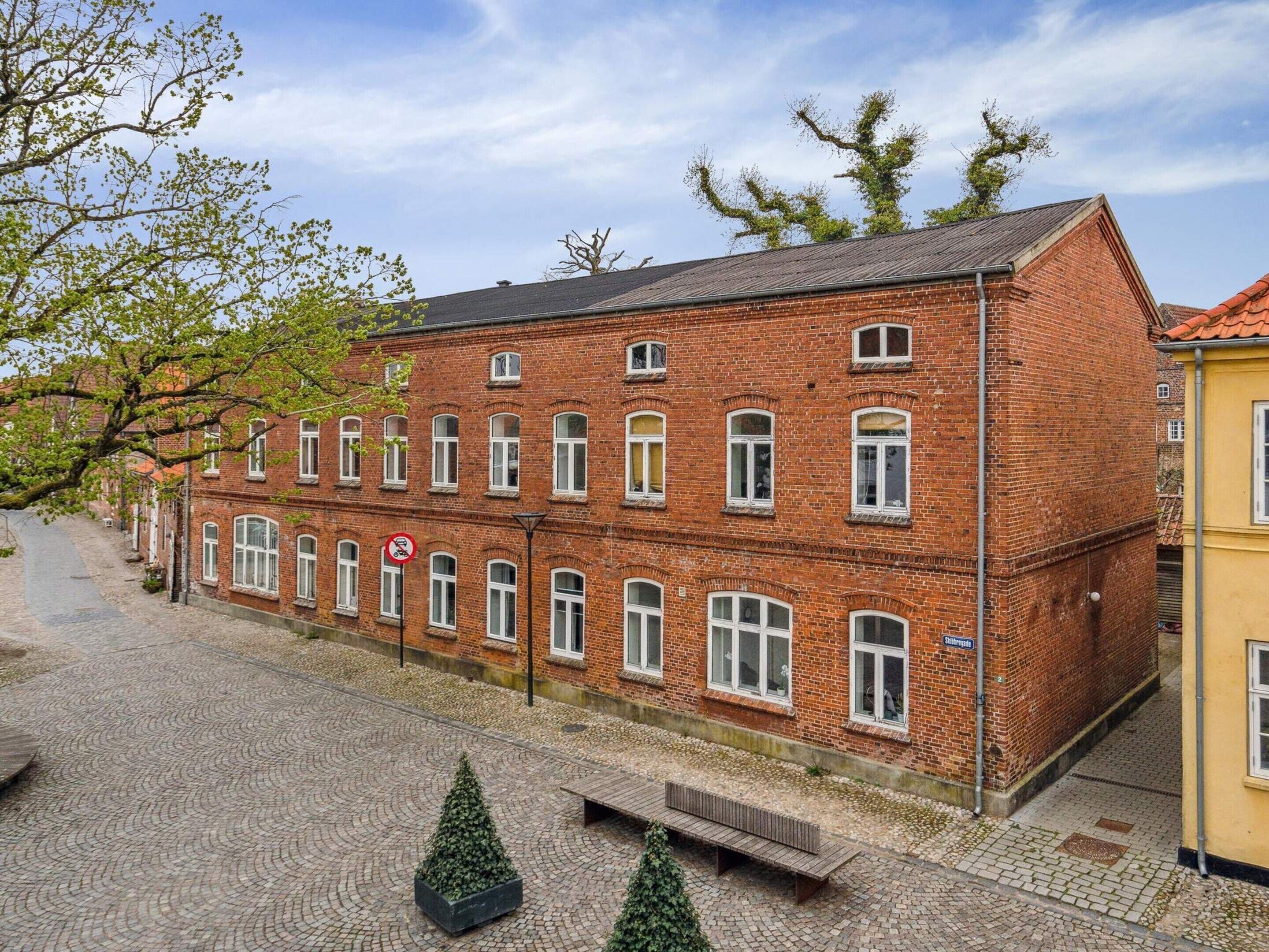 A red brick building with multiple windows, a paved courtyard, and a bench.
