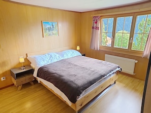Ein Schlafzimmer mit einem hölzernen Bett, einem Nachttisch mit Lampe, einem Fenster mit Blick ins Grüne und einem Bild an der Wand.