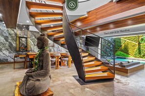 Ein modernes Interieur mit einer Wendeltreppe, einer Statue, einem Essbereich und Blick auf eine grüne Außenanlage.