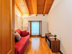Ein Zimmer mit Holzbalkendecke, einem roten Sofa, einem kleinen Tisch mit Pflanze und einer Tür, die auf einen Balkon führt.