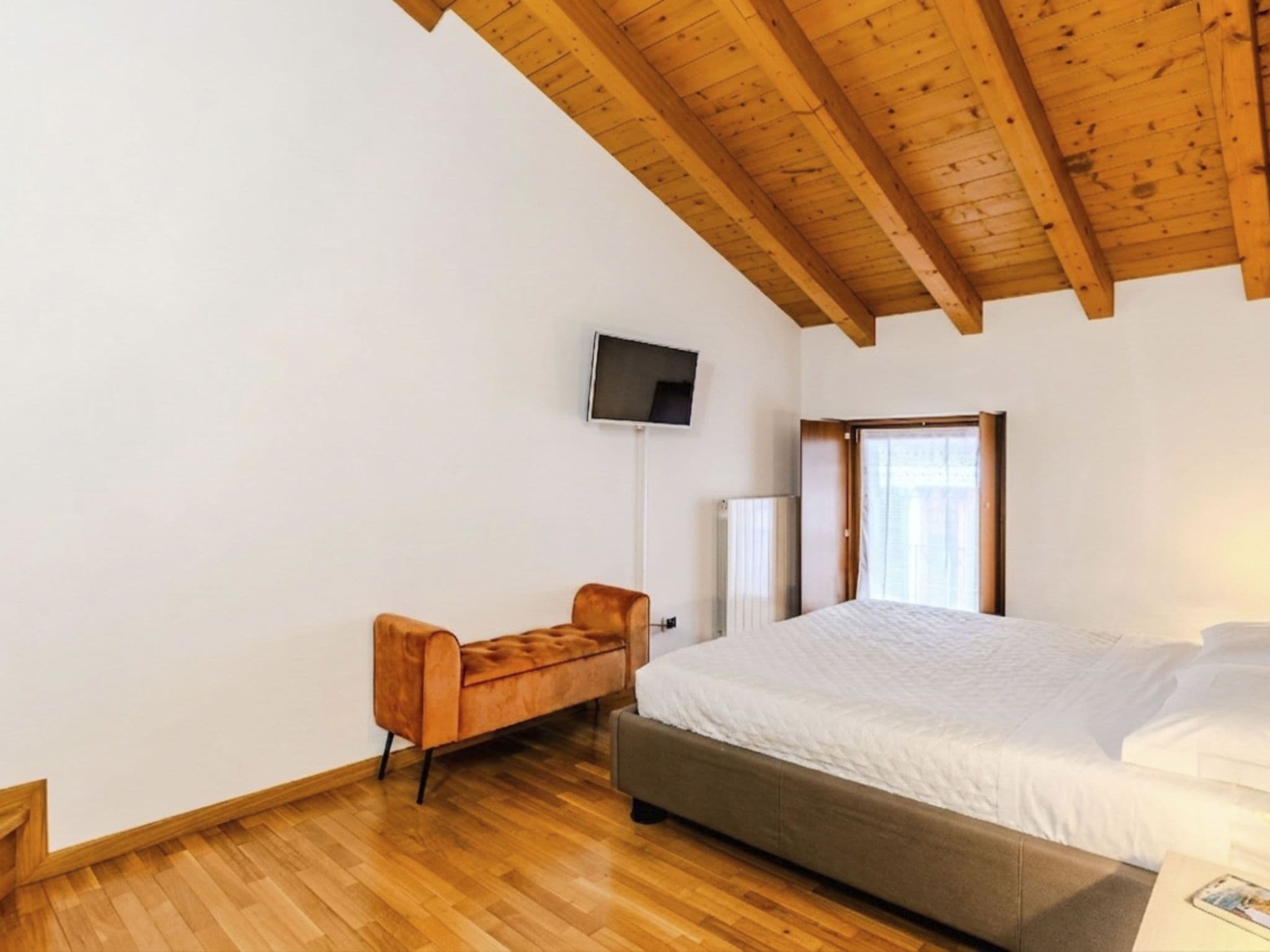 A bedroom with a wooden ceiling, a bed, a TV mounted on the wall, and a window with curtains.