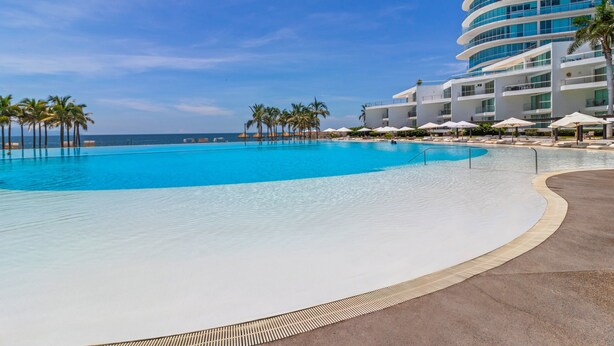 Ein großer Pool mit geschwungener Kante, umgeben von Palmen und einem modernen Gebäude mit Balkonen.